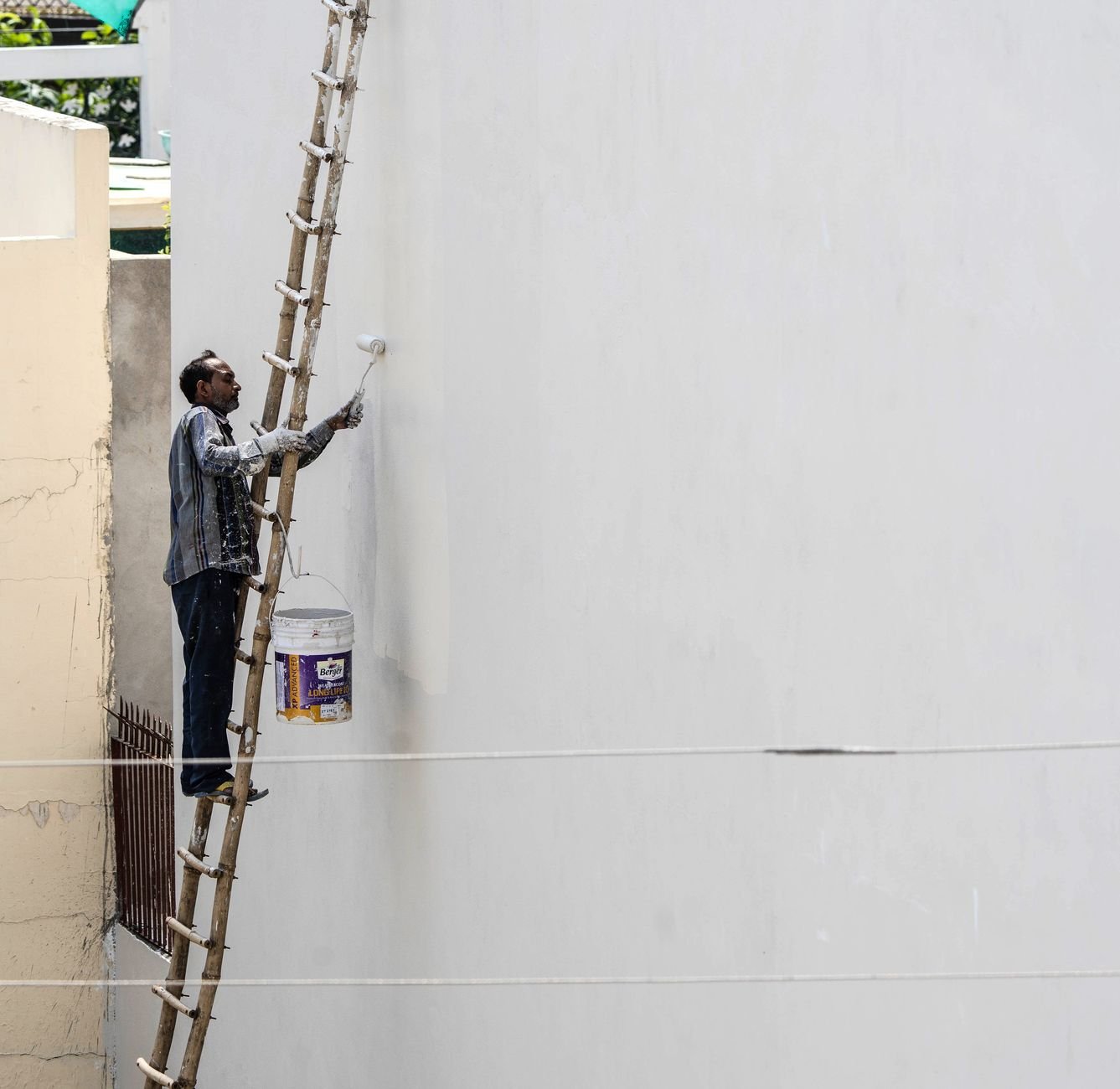 A man on a ladder painting a building's exterior wall with a roller brush outdoors.